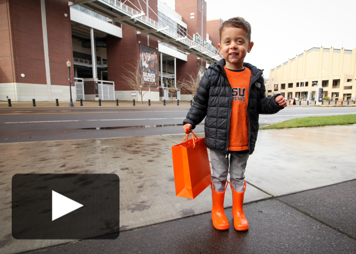 Young boy dressed in orange shirt, grey pants, black coat and orange rain boots holds an orange gift bag with Reser Stadium in the background and a play button place din lower lefthand corner
