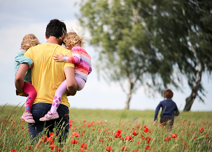 Photo of father carrying two young children through a poppy field while another child runs ahead