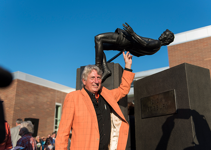 Dick Fosbury picured in orange sportcoat pointing to a sculpture of himself in the background at the dedication event