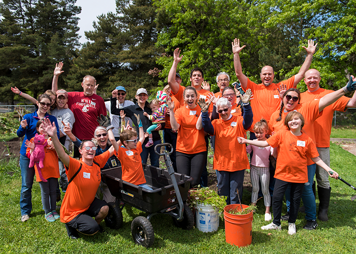 DOS volunteers at a gardening project wearing orange DOS shirts and posing for a photo with smiles and their hands in the air