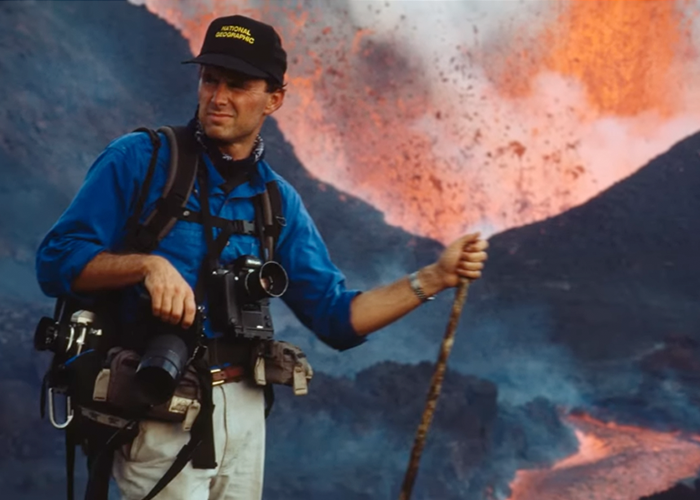 Screenshot from video showing Chris Johns with camera equipment and walking stick and lava shooting out from a volcano