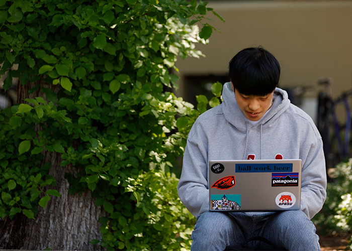 student smiling while sitting outside on sunny day and using a laptop