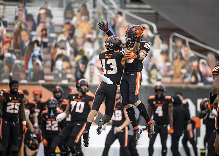 Two Beaver football players jump in air and chest bump on the field with other players and stands in the background