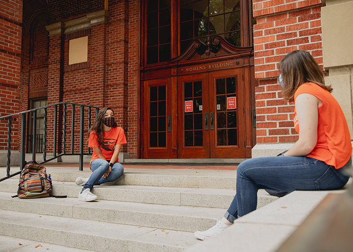 Photo of two students sitting in front of the Women's Building using masks and social distacing to hang out