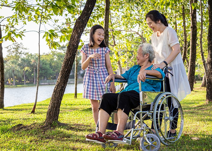 Photo of a child laughing with elder being pushed in a wheelchair by another adult in a sunny outdoor setting